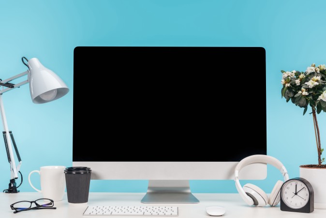 workplace with computer, lamp, cup, coffee to go, clock and flowerpot on white table on blue